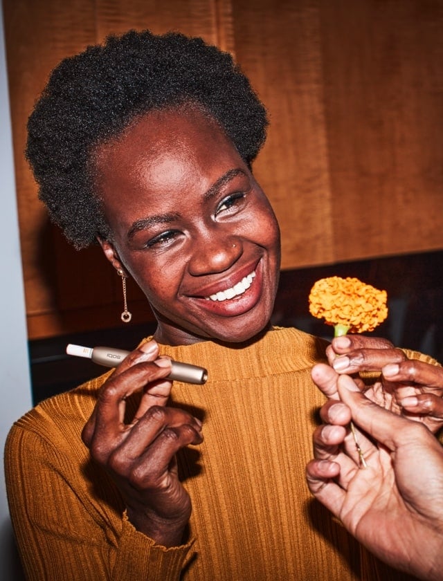 Girl smiling with an IQOS device and HEETS in one hand, receiving a yellow flower with the other